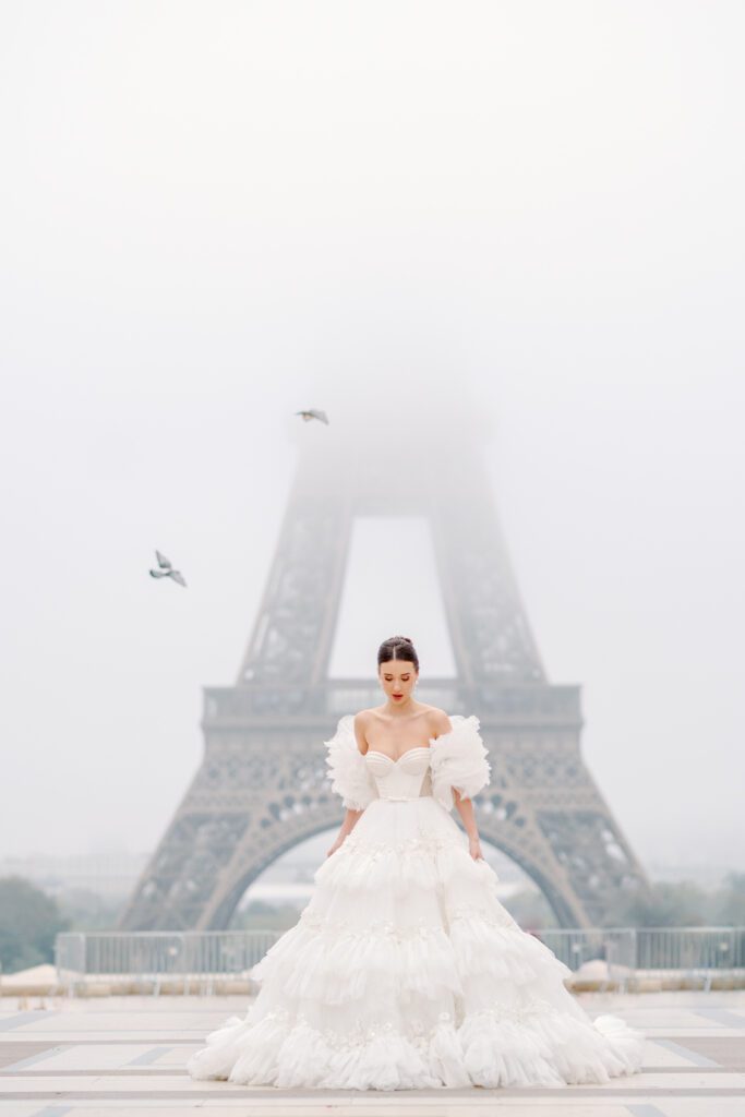 Paris pre-wedding session in front of the Eiffel Tower while it is covered in fog with bride in full train layered gown with separate off the shoulder full puffed sleeves and groom in a navy blue suit photographed by Italy destination photographer